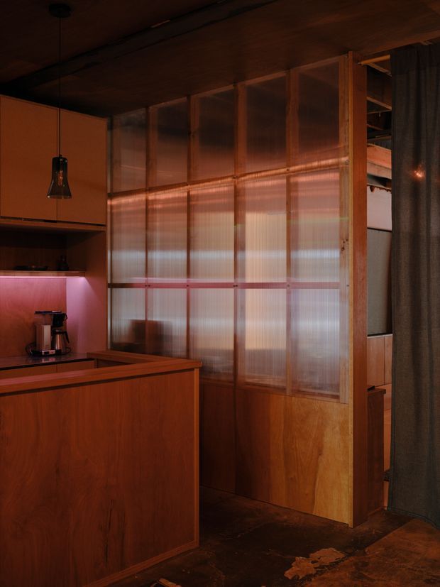 A dimly lit compact kitchen or kitchenette with warm timber cabinetry and a peninsula counter. A translucent corrugated-panel partition in timber framing separates the kitchen from an adjacent space, while a dark curtain partially screens an opening on the right. Coppery ambient lighting, pendant lighting, dark flooring, and rich wood surfaces create an intimate, moody interior.