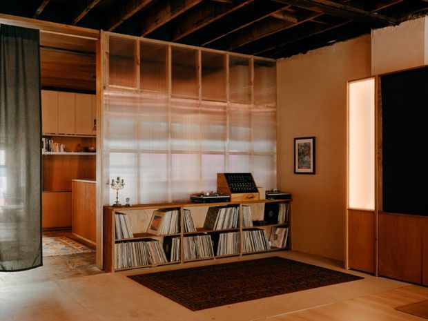 Warm, moody open-plan living room with a timber record-storage and media console, extensive vinyl collection, translucent ribbed-glass room divider, exposed dark timber ceiling structure, and a concealed kitchen or kitchenette visible beyond. Terracotta-toned walls, timber joinery, concrete-like flooring, and a dark patterned rug create a rich, vintage-inspired atmosphere.