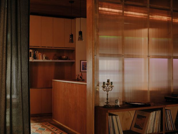 A warm, moody interior view of a compact mid-century-inspired living area and adjoining kitchen or bar zone. Rich timber cabinetry, shelving and a timber console dominate the space. Ribbed translucent glass partitions or sliding panels separate the foreground media/storage area from the rear joinery, while dark pendant lights, vinyl records and a patterned tiled floor add visual character.