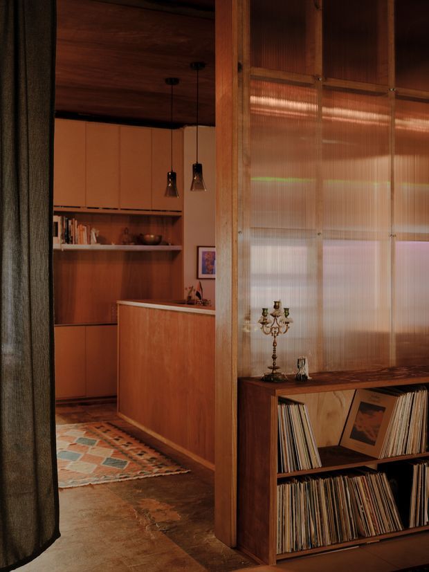 Warm, moody interior view looking through a doorway into a compact timber-lined kitchen. The kitchen features wood cabinetry, open shelving, a peninsula, dark flooring, and pendant lights. In the foreground, a timber record console with vinyl storage sits beneath a translucent ribbed glass or polycarbonate partition framed in timber. Rich brown wood tones, terracotta cabinetry, and vintage furnishings create a retro, intimate atmosphere.