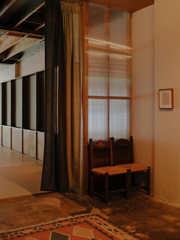 A warm, dimly lit interior corner with two dark timber chairs positioned beneath a tall translucent window framed in timber. Heavy dark curtains divide the seating area from an adjacent open space with exposed ceiling and structural elements and a series of dark partition panels. The room features earthy plaster walls, a timber ceiling, a worn dark floor, and a geometric patterned rug in terracotta, cream, and dark tones. A small framed artwork hangs on the wall.