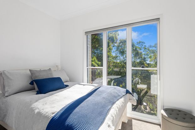 Bright, compact bedroom with white walls and bedding, a grey upholstered bedhead, and layered blue and white bedding. Full-height glazed doors open to a balcony or terrace with leafy outdoor views, bringing abundant natural light into the room. A small grey upholstered stool sits beside the bed.