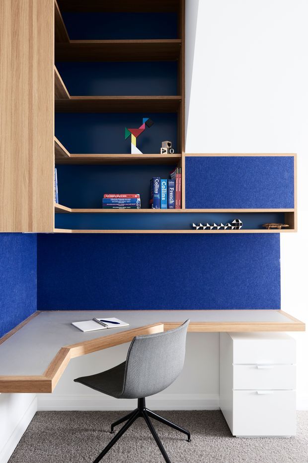 A compact built-in home office with an L-shaped light grey worktop edged in timber, white drawer cabinetry, and a grey upholstered swivel chair. Above the desk is a wall-mounted timber shelving unit with deep navy-blue backing and a matching navy wall panel, holding books and small decorative objects. The space is bright, tailored, and contemporary.
