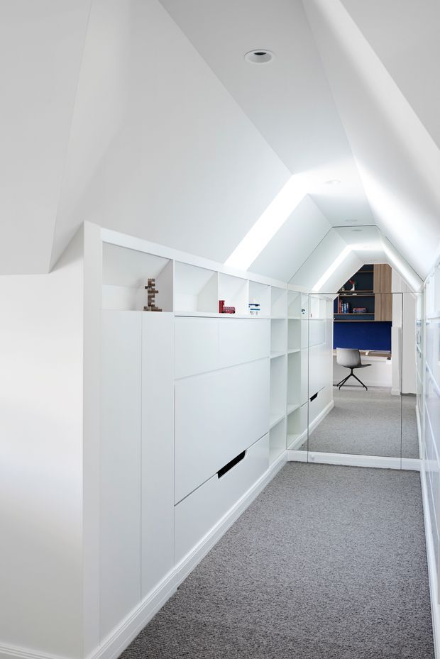 A bright, narrow attic-style home office or study corridor with a sharply sloped white ceiling, recessed downlights, continuous integrated white cabinetry and open shelving, and grey carpet flooring. A mirrored wall reflects a compact desk area at the far end with a white swivel chair, blue wall backing, and timber-toned shelving. The space is highly streamlined and built-in, with strong linear LED lighting following the ceiling angles.
