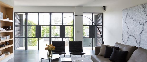 Bright contemporary living room with black-framed floor-to-ceiling glazing and louvred side panels, built-in open shelving, pale flooring, a large curved arc floor lamp, dark lounge chairs, and a grey sectional sofa. A large abstract textured artwork forms a feature wall, while greenery is visible beyond the glazing.