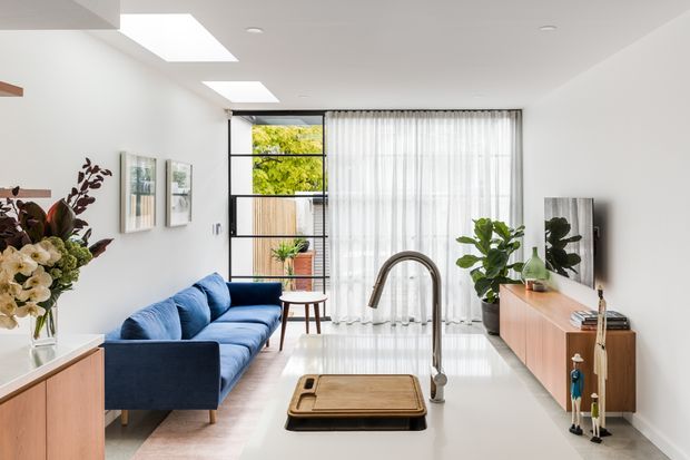 Bright contemporary open-plan living room and kitchen with a long white island, integrated curved tap, blue sofa, timber media console, indoor plants, sheer curtains, and floor-to-ceiling glazing. The room has polished light flooring, white walls and ceiling, black-framed glazing, skylights, and abundant natural light.