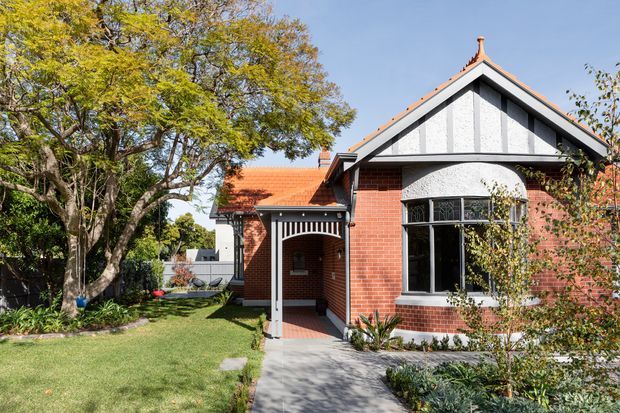 Exterior view of a heritage-style brick house with a prominent front-facing gabled roof, terracotta roof tiles, decorative white gable infill and timber detailing, black-framed windows, and a covered entry porch. The home is set within a landscaped suburban garden with mature trees and a paved front path.