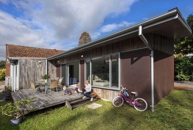 Exterior view of a compact modern single-storey dwelling with a low-pitched roof, timber-clad walls, dark brown panels, and large glazed doors and windows. A raised timber deck with outdoor dining furniture fronts the building, while a bicycle and landscaped garden occupy the foreground. An older tiled-roof timber structure is attached or adjacent at the left.