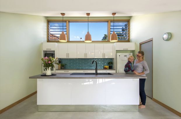 Bright contemporary kitchen viewed straight on, featuring a large white island with integrated sink, glossy white wall cabinetry, a dark benchtop, teal rectangular tile splashback, timber-framed horizontal windows, and three pendant lights. A parent and child stand at the right side of the island. The room has pale green walls, concrete-look flooring, and a clean, family-oriented layout.