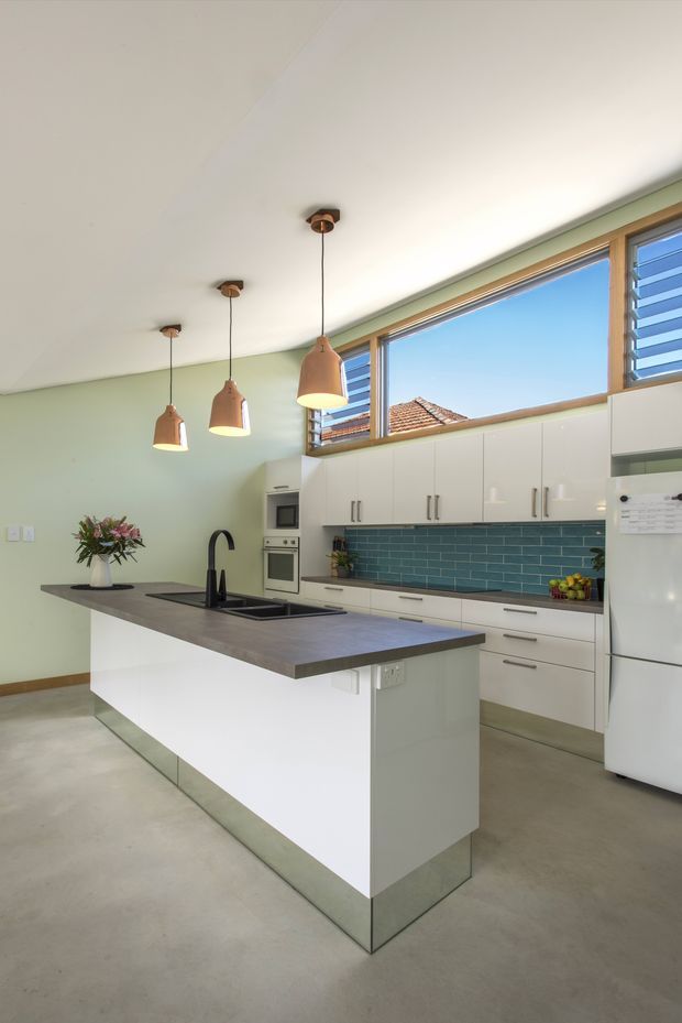 Bright contemporary kitchen with a large white island, dark grey benchtop, black undermount sink and tap, white handle cabinetry, teal-blue subway tile splashback, polished concrete floor, and high clerestory-style louvred windows. Three copper pendant lights hang above the island, with integrated cooking appliances and a refrigerator along the rear wall.