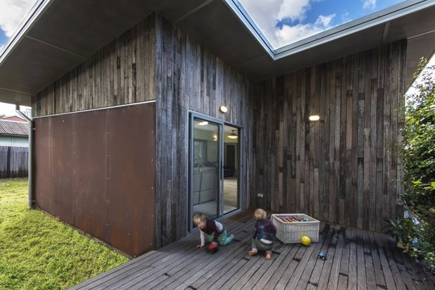 Exterior view of a compact contemporary house or extension clad in dark weathered vertical timber boards. A recessed entry opens onto a timber deck, with a large rust-brown metal panel door and simple exterior wall lights. The roof has a strong angular form with a glazed skylight or roof opening, and children are playing on the deck.