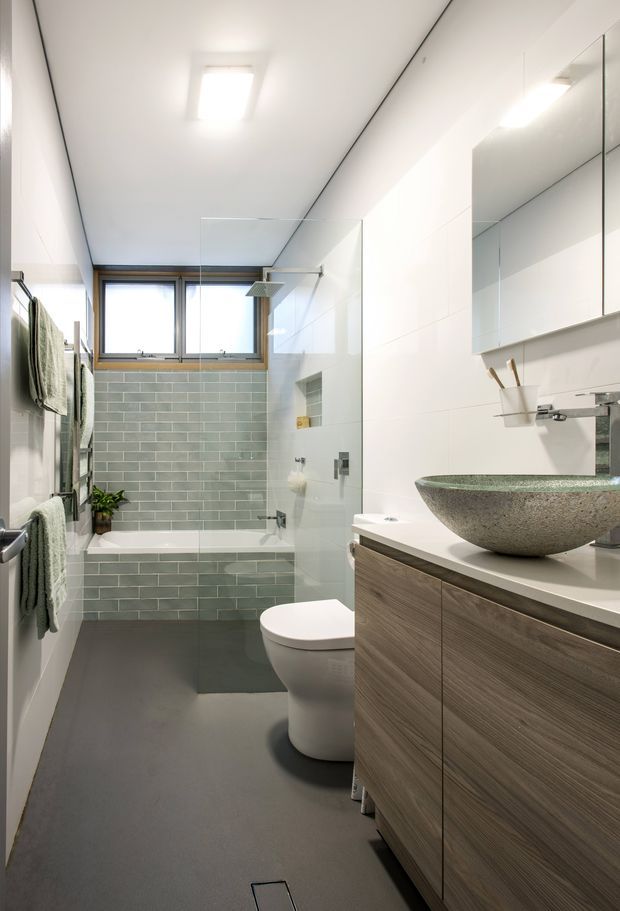 A bright, narrow contemporary bathroom with a bathtub and glass shower screen at the rear, a wall-hung toilet, and a long timber-grain vanity with a large round stone vessel basin. Pale green-grey rectangular wall tiles surround the bath, while the remaining walls and ceiling are white. A dark grey continuous floor, mirrored wall cabinets, chrome fixtures, recessed shelving, and towel rails complete the clean modern fit-out.