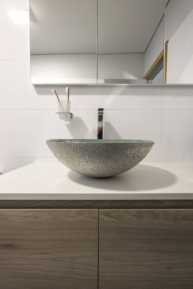 Close-up of a contemporary bathroom vanity featuring a textured grey-green stone vessel basin, chrome wall-mounted tap, white tiled wall, mirrored storage cabinet, white benchtop, and dark timber-grain vanity cabinetry. A small wall-mounted toothbrush holder is visible beside the basin.