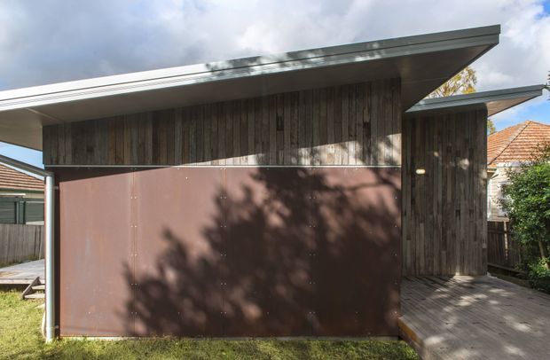 Exterior view of a compact contemporary dwelling or extension with a strongly projecting asymmetrical roof, vertical weathered timber cladding, and a large dark brown metal-clad wall or garage door. A timber decked entry walkway leads into a recessed opening. The building sits in a suburban residential setting, with neighbouring rooflines and fencing visible around it.