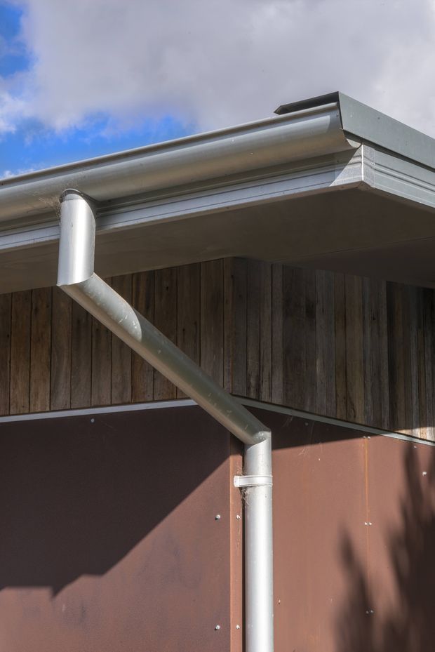 Close-up exterior detail of a contemporary building corner, showing a flat roof edge with metal guttering and downpipes, timber vertical wall cladding, and a dark reddish-brown metal wall panel beneath. Bright daylight and blue sky are visible above.