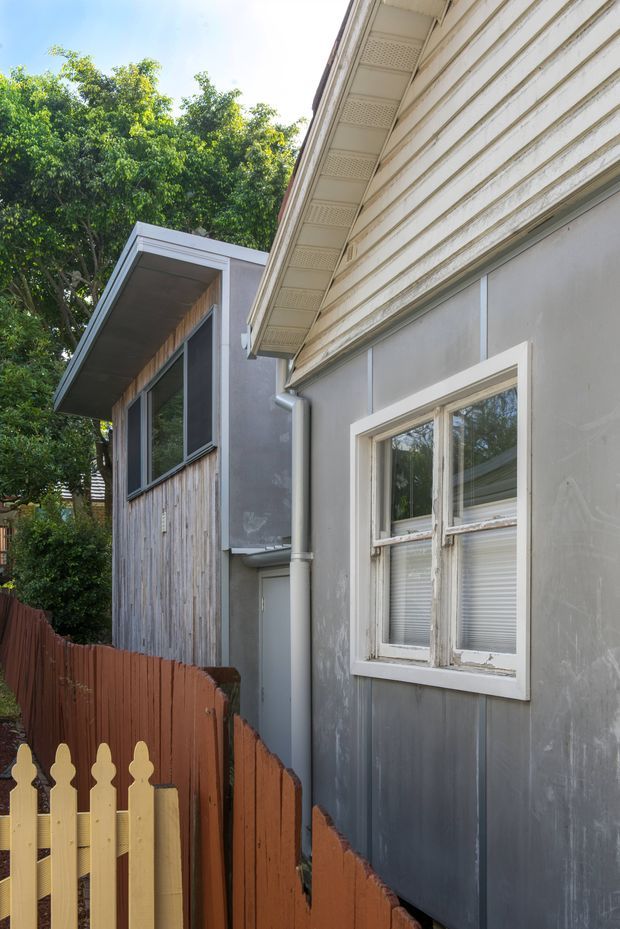 A narrow residential side passage between two adjoining building volumes. The foreground facade has grey painted wall cladding, a white-trimmed window, horizontal weatherboard-style detailing near the eaves, perforated soffit lining, and a downpipe. A smaller modern extension with dark-framed glazing and vertical timber cladding is visible beyond a red-brown and pale timber picket fence. Mature trees surround the property.