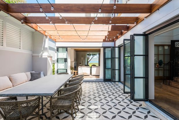 A bright, covered indoor-outdoor entertaining area with a long dining table and woven chairs, patterned geometric floor tiles, timber pergola beams, string lights, louvred wall panels, and large black-framed folding glass doors opening toward a landscaped outlook. A living or circulation space is visible through the open doors on the right, with another seating area beyond.