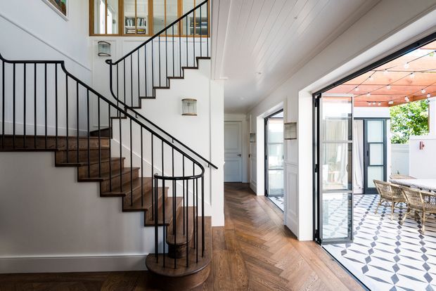 Bright, refined residential hallway centred on a sculptural staircase with dark timber treads, slim black metal balustrades and white walls. Herringbone timber flooring continues through the circulation space toward large glazed doors opening onto a light-filled tiled outdoor terrace or courtyard with dining furniture. A white-lined ceiling and generous glazing create an airy, connected indoor–outdoor feel.
