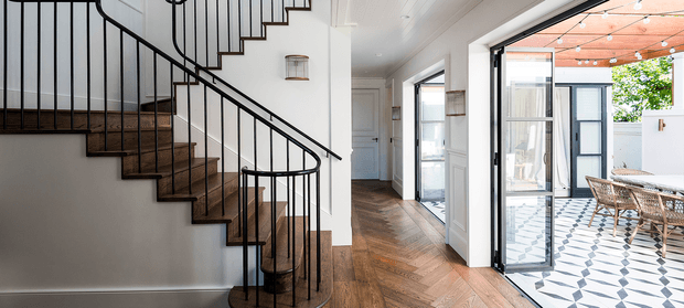 Bright contemporary residential hallway and stairway with a sculptural curved staircase, dark timber treads, slim black metal balustrade, white walls, and warm herringbone timber flooring. The hallway opens through black-framed glazing to a covered outdoor dining courtyard featuring patterned floor tiles, timber ceiling lining, and wicker furniture.