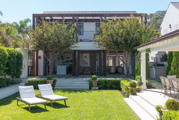 Bright landscaped courtyard garden viewed toward a two-storey contemporary residence. The home has a pale rendered facade, dark timber pergola and balcony framing, extensive glazed openings, and a central covered outdoor living area. A manicured lawn, clipped hedges, potted plants, sun loungers, and a side terrace with outdoor dining create a refined resort-like setting.