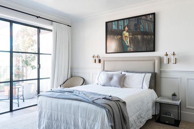 Bright, refined bedroom with a large upholstered bed, white quilted bedding, layered grey throws, paneled white walls, black-framed floor-to-ceiling glazed doors, and a framed artwork above the bed. Slim bedside furniture and wall-mounted decorative lights create a polished contemporary look.