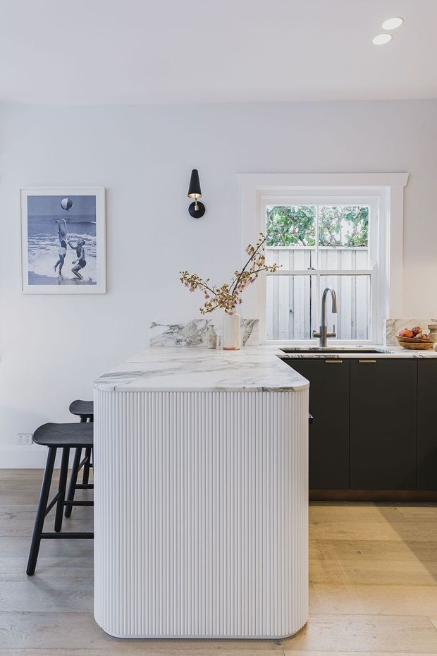 Bright contemporary kitchen featuring a large white marble island with rounded ends and vertically fluted cabinetry, paired with matte black lower cabinets and a dark sink. A timber-framed window, pale walls, light timber flooring, pendant-style wall lighting, and restrained artwork create a refined, modern interior.