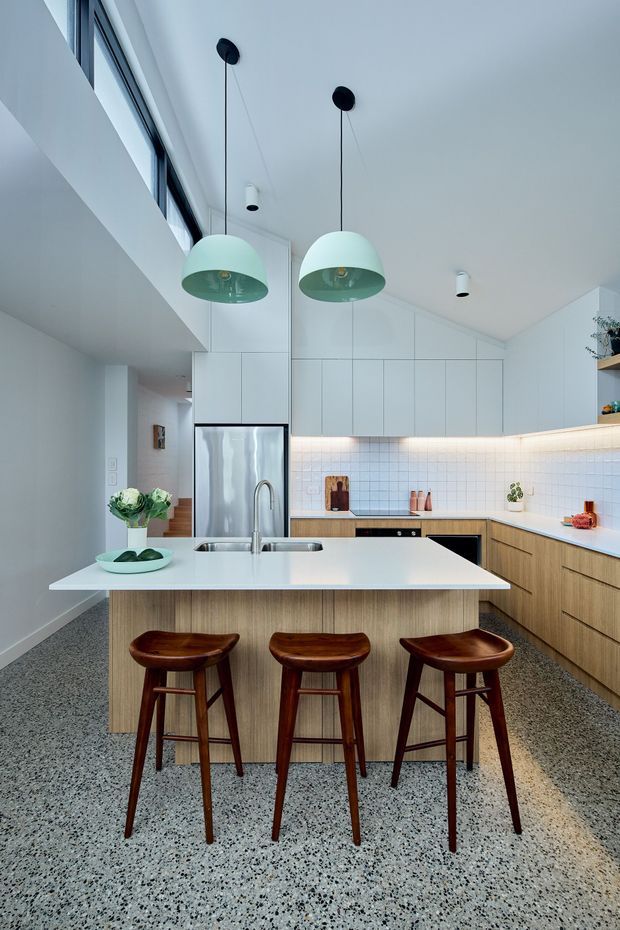 Bright contemporary kitchen with a large island and three timber bar stools. The room features white full-height cabinetry, warm timber base cabinets, a pale tiled splashback, integrated stainless-steel appliances, a white island benchtop, terrazzo-style flooring, mint-green pendant lights, and a high sloping ceiling with clerestory glazing.