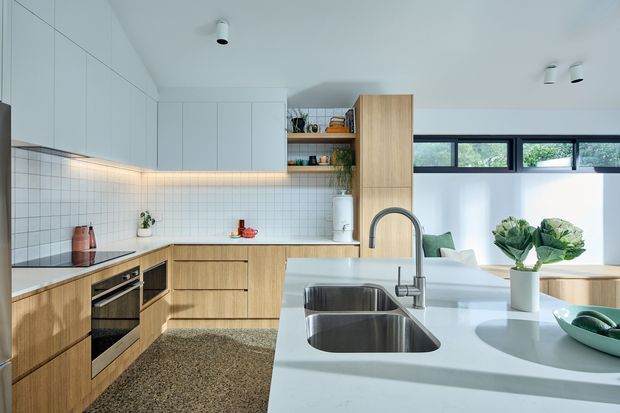 Bright contemporary kitchen with light timber cabinetry, a white tiled splashback, white benchtops, integrated appliances, open shelving, and a large island with a double stainless-steel sink. A dining or living area is visible beyond the kitchen, with high horizontal windows providing natural light.