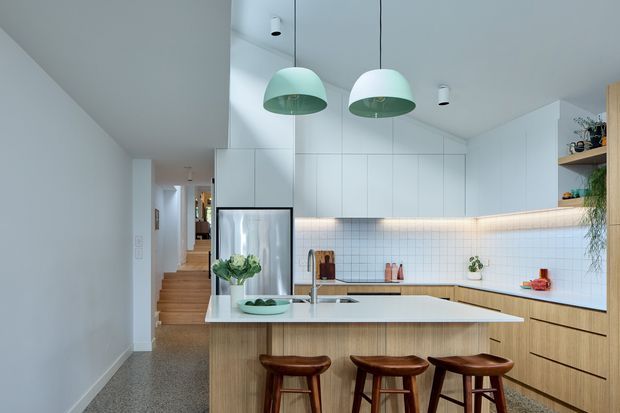 Bright, contemporary open-plan kitchen viewed from the living or circulation area. A large timber-clad island with three wooden stools faces a white stone-look benchtop, with pale timber lower cabinetry, white upper cabinets, a small square-tile splashback, integrated refrigerator, and mint-green pendant lights. A hallway and timber stairway are visible beyond the kitchen.