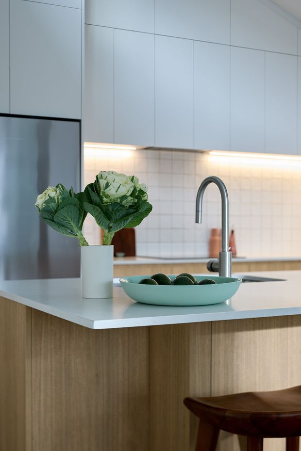 Close-up view of a contemporary kitchen island with a pale stone-look benchtop, vertical light timber cabinetry, white flat-panel overhead cabinets, and a light tiled splashback. A stainless-steel refrigerator is visible at left, while a curved stainless-steel mixer tap and inset sink sit behind a turquoise bowl. Under-cabinet lighting illuminates the work area, and a timber stool is partially visible in the foreground. The styling is clean, restrained, and modern with natural timber accents.