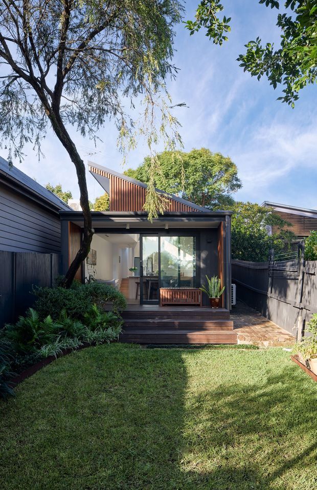 Exterior view of a compact contemporary backyard dwelling or extension, set behind a landscaped lawn. The building has dark horizontal cladding, a steep asymmetrical roof form with vertical timber detailing, large glazed openings, and a small raised timber porch with built-in bench seating. Brick paving, planting, boundary fencing, and mature trees frame the sunny garden setting.