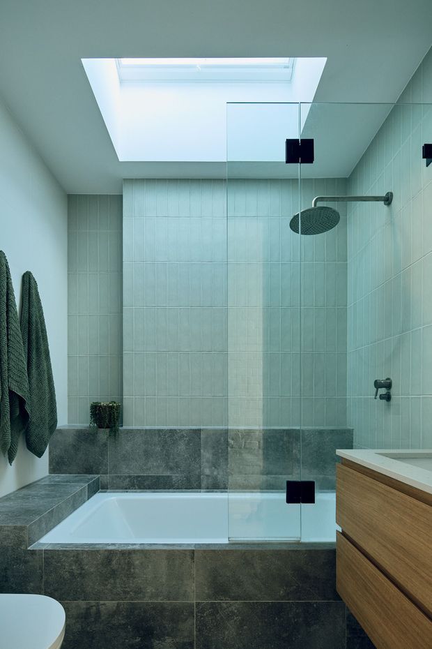 A compact contemporary bathroom featuring a tiled bath and shower area behind a frameless glass screen, a large skylight, wall-mounted rainfall shower, bathtub, dark stone-look tiles, pale green vertically patterned wall tiles, and a timber vanity with a light benchtop. Dark textured towels hang on the left wall.