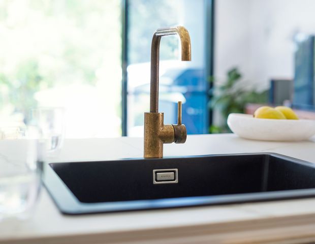 Close-up view of a modern kitchen sink with a deep matte-black undermount or flush-mounted basin, white benchtop, and a tall brushed bronze or brass gooseneck mixer tap. Bright floor-to-ceiling glazing and soft greenery are visible in the background, with a white fruit bowl on the counter. The image has a clean, contemporary, lightly warm and airy aesthetic.