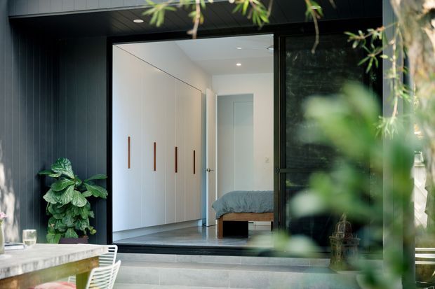 View from an outdoor dining area toward a modern home entry, with dark vertical exterior cladding framing a large glazed opening. Inside, a bright minimalist bedroom is visible, featuring full-height white built-in wardrobes with slim timber handles, a grey bed, pale walls, and a polished grey floor. Steps lead up to the entry, with lush planting partially screening the view.