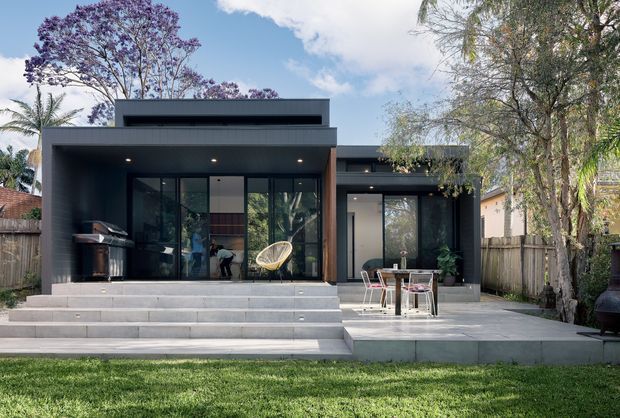 A modern single-storey residential house viewed from the rear garden, with dark charcoal cladding, broad floor-to-ceiling glazing, a flat roof, and a stepped concrete terrace. The open outdoor living area includes a built-in barbecue, dining furniture, and a small seating nook. A lawn, mature trees, and neighbouring residential fencing surround the home.