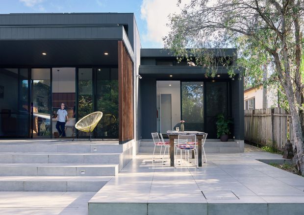 Exterior view of a contemporary single-storey home with dark cladding, broad floor-to-ceiling glazing, a covered terrace, and stepped pale paving. The house opens onto an outdoor dining area and raised patio, with timber screening and established trees along the boundary.