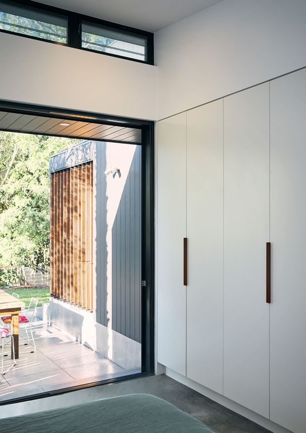 A bright minimalist interior corner with full-height white built-in cabinetry and slim dark timber handles. Black-framed glazing and a high clerestory window open to a sunny outdoor patio, while a partial upholstered seating surface appears in the foreground. The space has clean linear detailing, a polished concrete floor, and a restrained neutral palette with warm timber visible outside.