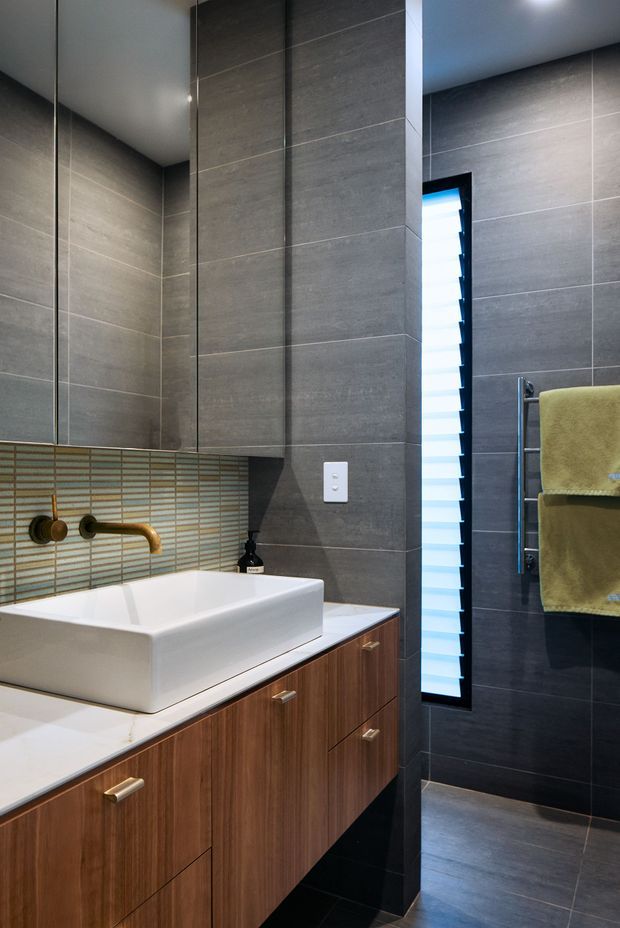 A contemporary bathroom featuring a floating timber vanity with a white vessel basin, brass wall-mounted tapware, large grey tiled walls, a narrow louvred window, mirrored cabinetry, and a heated towel rail with olive towels. The space has clean linear detailing, dark flooring, and integrated storage.