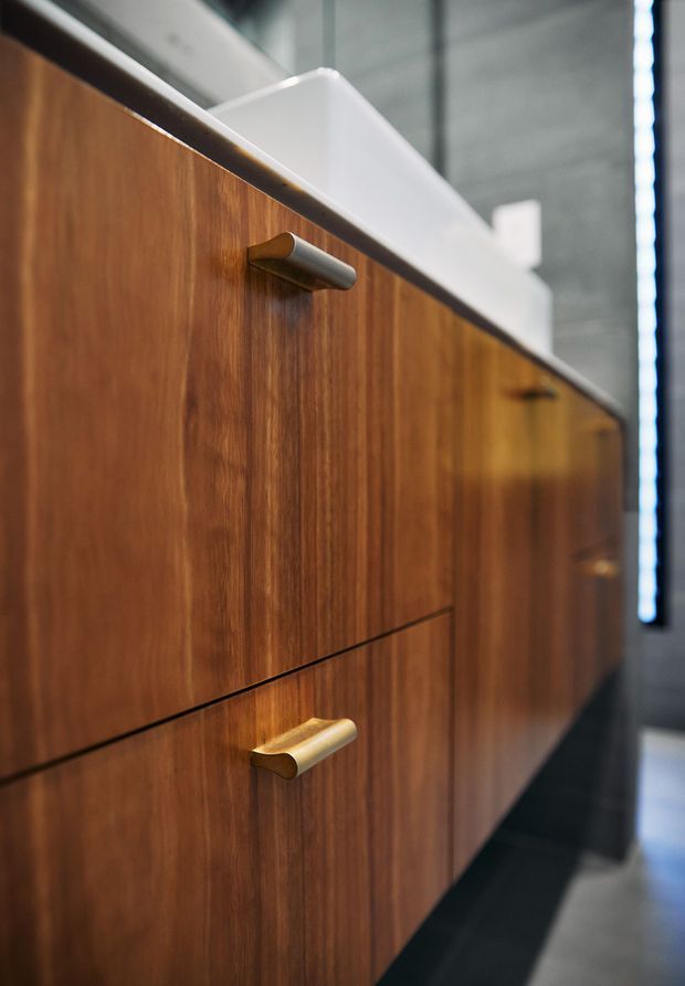 Close-up view of a contemporary vanity or built-in cabinet with rich medium-to-dark timber veneer fronts, slim horizontal drawers, and contrasting cylindrical metal pulls in brushed silver and brass-gold finishes. A white integrated basin or benchtop is visible above the cabinetry, with a dark floor and grey architectural surfaces in the background. The image emphasises refined joinery and hardware rather than the full room.