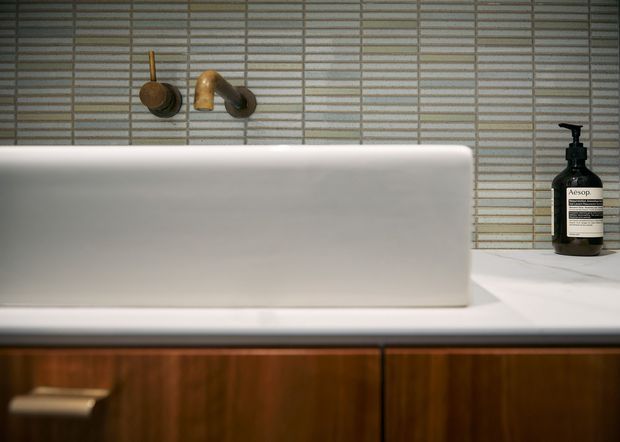 Close-up of a refined bathroom vanity featuring a freestanding white rectangular basin, wall-mounted aged brass tapware, narrow horizontal tiled splashback, white benchtop, and dark timber cabinetry with brass handles. An Aesop soap dispenser sits at the right edge. The composition is minimalist and warm, with a restrained neutral palette and rich wood joinery.