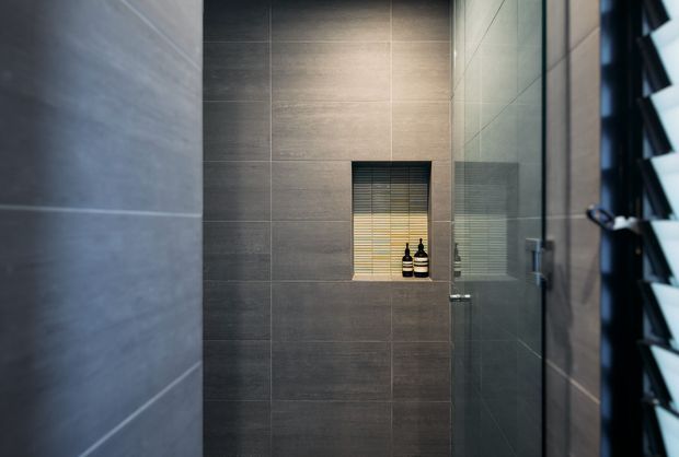 A compact, modern tiled bathroom or shower enclosure viewed through a partially open glass door. Large-format dark grey wall tiles cover the walls, with a recessed wall niche containing small bottles. A louvred window on the right provides filtered daylight, and the space has a clean, restrained contemporary appearance.