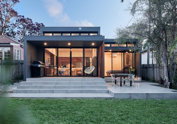 Rear exterior view of a contemporary single-storey home extension with a dark framed, extensively glazed living area opening onto a broad concrete terrace and stepped entry platform. Warm interior lighting reveals a dining/living space, while an outdoor table and chairs occupy the terrace. The addition contrasts with the neighbouring traditional homes and is framed by established trees and lawn.