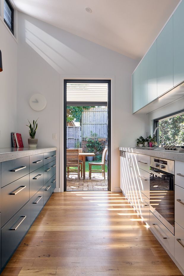 Bright, narrow galley kitchen with pale blue-gray and white built-in cabinetry on both sides, light stone-look benchtops, stainless steel hardware and appliances, and warm timber flooring. A glazed door at the end opens to a small paved outdoor dining courtyard, while strong sunlight creates linear shadows across the floor and cabinetry. The room has a high sloping ceiling and a clean, contemporary feel.