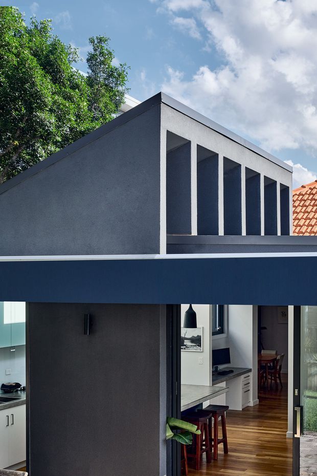 Exterior view of a contemporary two-storey residential extension with a dark navy horizontal upper fascia, charcoal rendered walls, and a distinctive row of deep vertical openings beneath a sloping roof edge. Large glazed openings reveal a bright kitchen and dining area inside, with timber flooring, white cabinetry, and dark stools. A red tiled roof from the adjoining building is visible at right, with mature greenery and blue sky in the background.