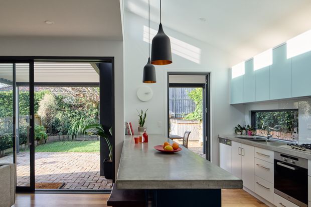 A bright contemporary kitchen viewed from the island, with a long concrete-look island benchtop, white lower cabinetry, pale blue-grey upper cabinets, stainless-steel appliances, and black pendant lights. Full-height glazing and a sliding glass door connect the kitchen to a landscaped paved courtyard. The room has a high ceiling and abundant natural light.