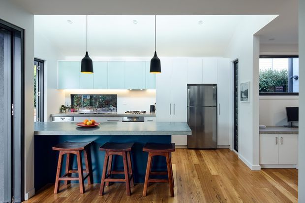 Bright contemporary kitchen with a large stone island and three timber bar stools, white full-height cabinetry, stainless-steel appliances, pale blue glass splashback, timber flooring, dark-framed glazing, and two black pendant lights. A built-in study nook is visible to the right.