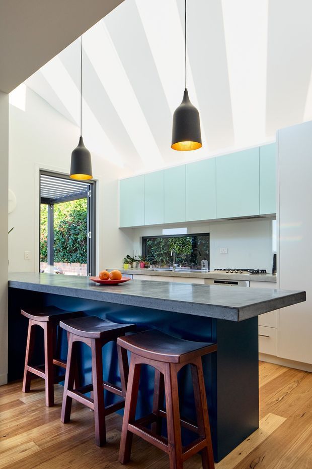 Bright contemporary kitchen featuring a large dark island with a grey stone benchtop and seating for three, pale blue-green full-height cabinetry, white walls, timber flooring, black pendant lights, and a high angled ceiling with skylights or clerestory glazing. An open glazed door connects to an outdoor area.