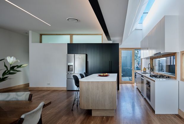 A bright contemporary open-plan kitchen and dining area with a large light-wood island, integrated stainless-steel appliances, white cabinetry, and a dark vertically panelled feature wall. Timber flooring extends through the space, with a glazed door and skylight bringing in natural light. A dining table and chairs are visible in the foreground.