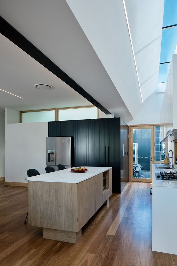 Contemporary kitchen with a large light-wood island and white stone benchtop, black vertically grooved feature cabinetry, stainless-steel refrigerator, white perimeter cabinetry, and dark timber flooring. The space has high ceilings, clerestory windows, a skylit void, and glazed timber-framed doors leading to an outdoor area. Integrated linear lighting and restrained detailing create a clean, refined interior.