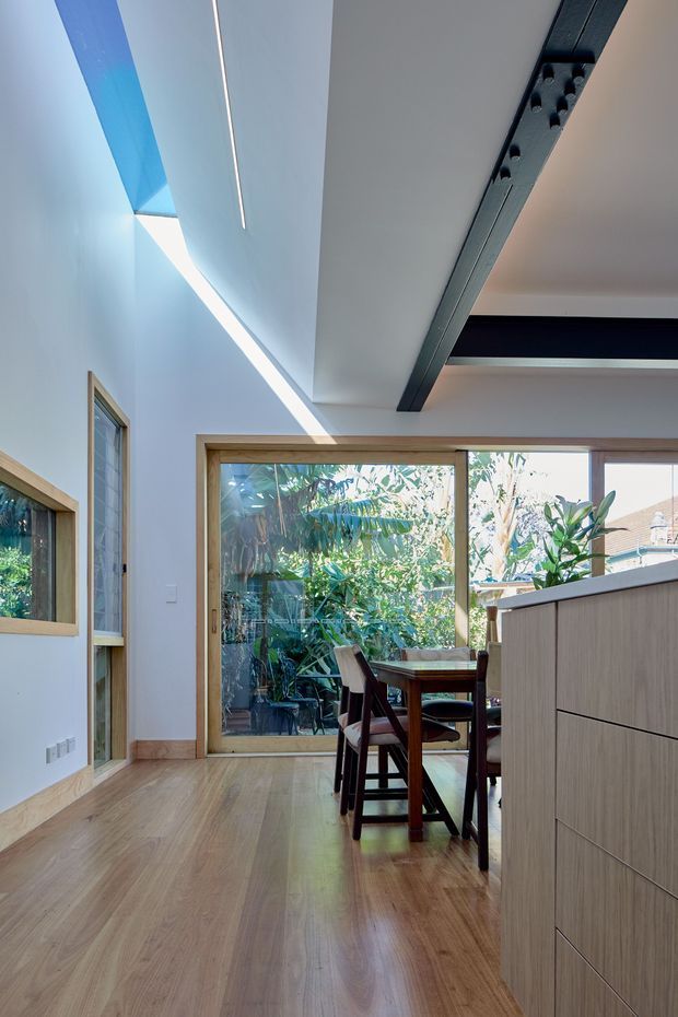 Bright contemporary open-plan interior viewed from a kitchen or circulation area toward a compact dining space. The room features warm timber-look flooring and window frames, pale walls, extensive glazing to a leafy outdoor area, light-wood built-in cabinetry in the foreground, and a dramatic exposed dark structural beam across the ceiling. Linear skylight or clerestory glazing introduces strong daylight.